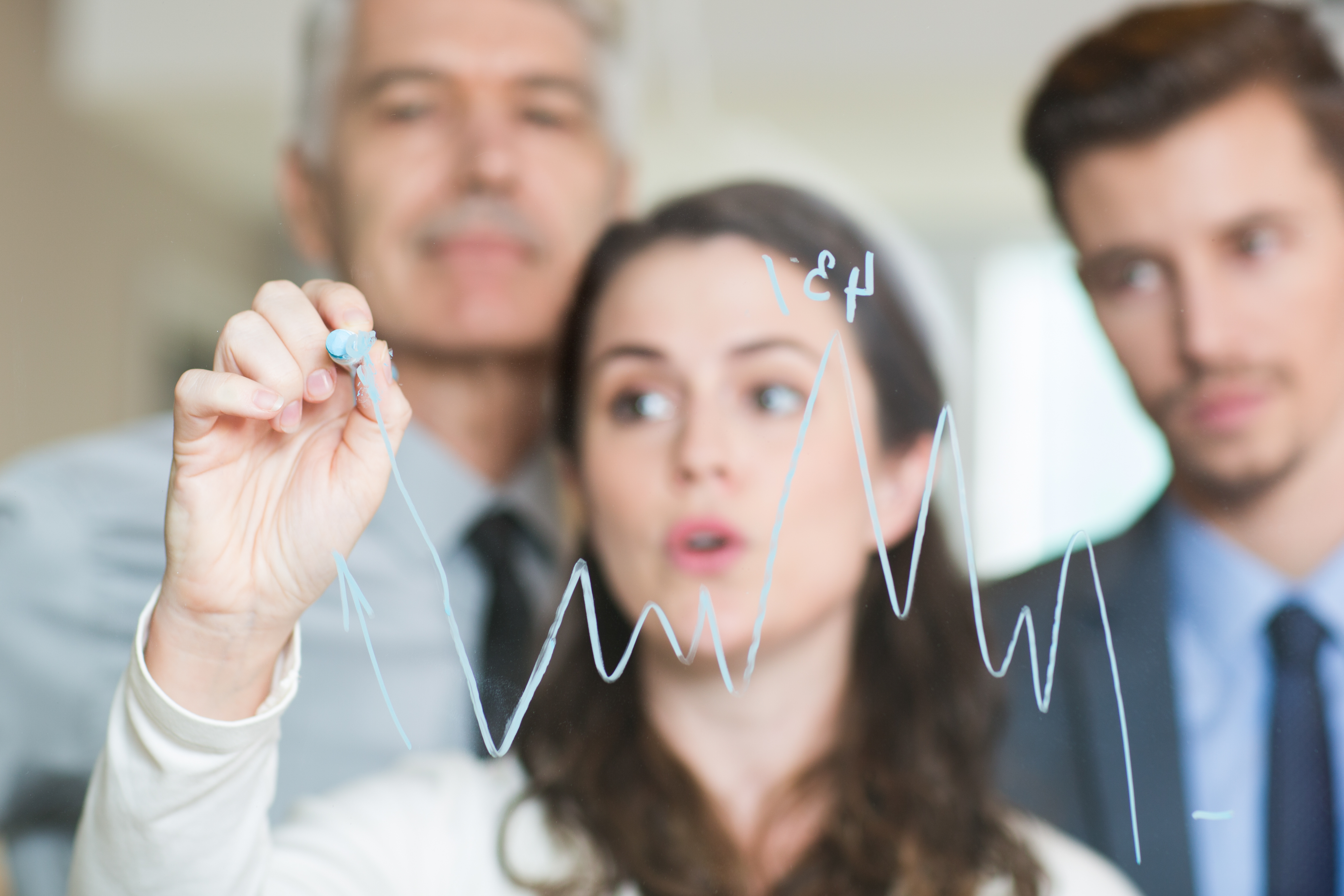 Closeup of graph drawn on transparent glass by blurred adult businesswoman standing behind, two young and senior colleagues with blurry office interior in background